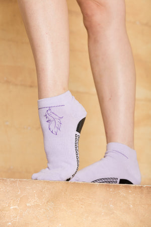 Profile close up of woman wearing lavender ankle grip socks accented with dark purple foil in the shape of a beaded anklet with a feather hanging from the chain. The chain extends from ankle to ankle on the front side of the foot.