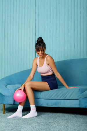 Woman sitting on a blue couch wearing white "Too Glam to Give a Damn" crew socks with a pink pilates ball, against a teal wall.