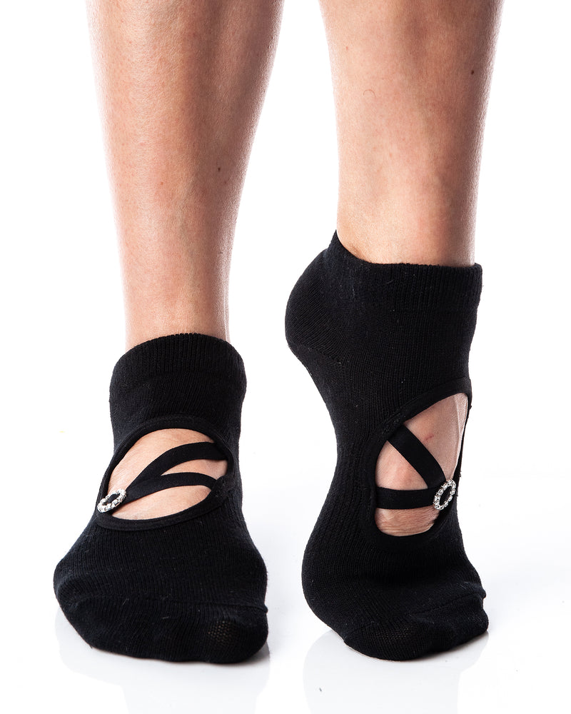 Black ankle height grip socks with a circular opening on the top of the foot. A diagonal strap connects with a horizontal strap across the circular opening, and a circular strap adjuster, accented with rhinestones, sits on the straps.