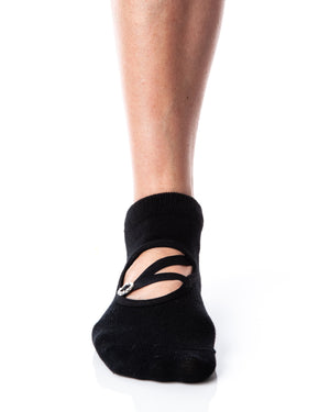 Detail shot of a single foot wearing black ankle height grip socks with a circular opening on the top of the foot. A diagonal strap connects with a horizontal strap across the circular opening, and a circular strap adjuster, accented with rhinestones, sits on the straps.