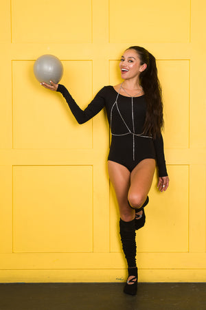Woman leaning against a yellow wall, holding a gray pilates ball while wearing ankle height grip socks with a circular opening on the top of the foot. A diagonal strap connects with a horizontal strap across the circular opening, and a circular strap adjuster, accented with rhinestones, sits on the straps.
